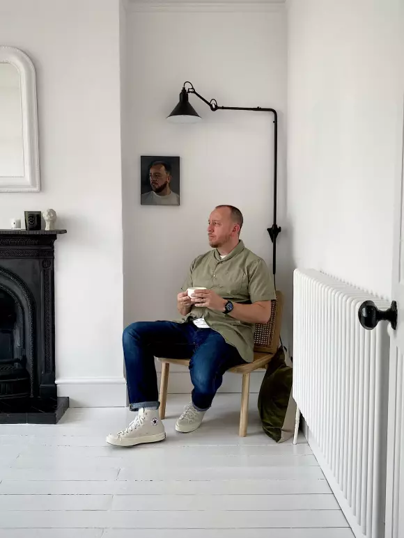 an image of a man sat in a chair next to a radiator holding a cup of tea in his hands