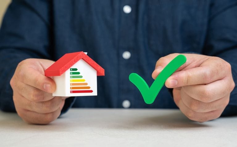 Close-up image of a man in a navy blue shirt holding a green tick next to a concept small white house with red roof displaying energy ratings