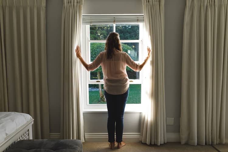 a woman opening the curtains in her bedroom to allow the sunlight to heat the space