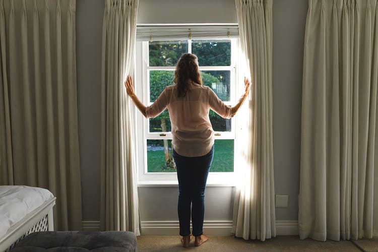 a woman opening the curtains in her bedroom to allow the sunlight to heat the space a woman opening the curtains in her bedroom to allow the sunlight to heat the space