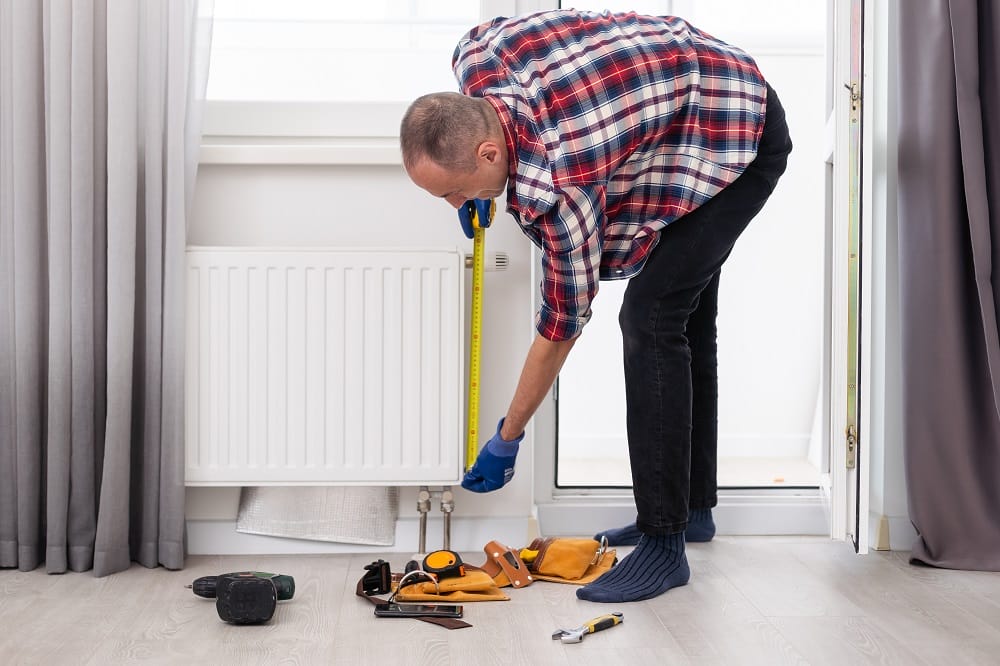 Man in gloves measuring a radiator for installation beneath a window with an array of tools