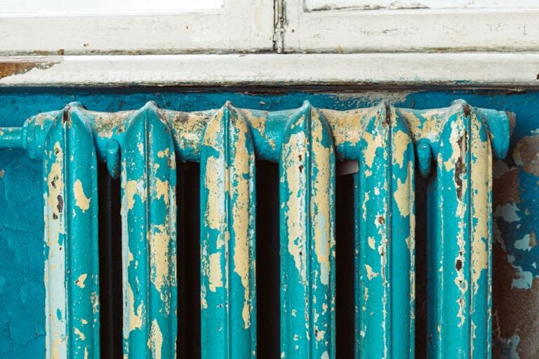 Close-up image of old cast iron radiator with peeling turquoise paint beneath weathered window sill