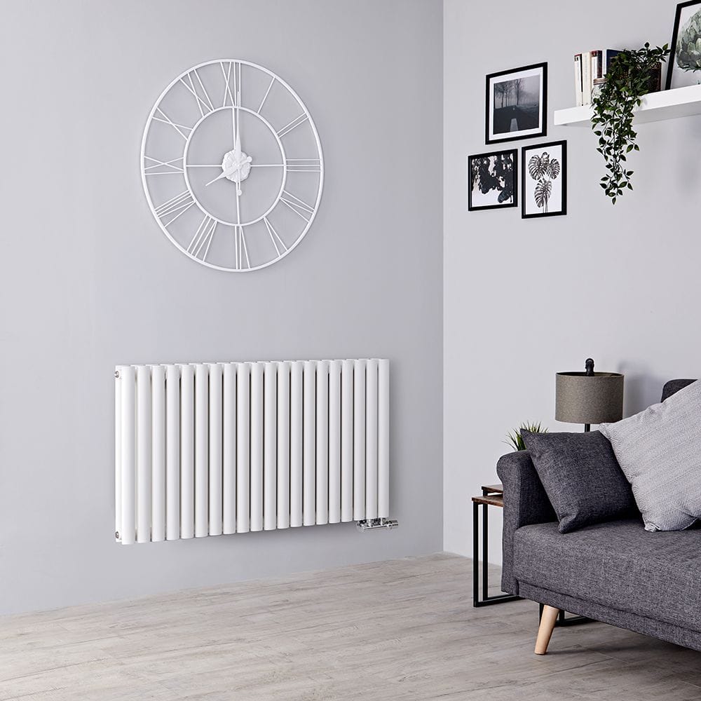 Milano Aruba Flow white horizontal side connection designer radiator on white wall beneath large white clock and nearby grey settee under shelf and multiple framed art pictures