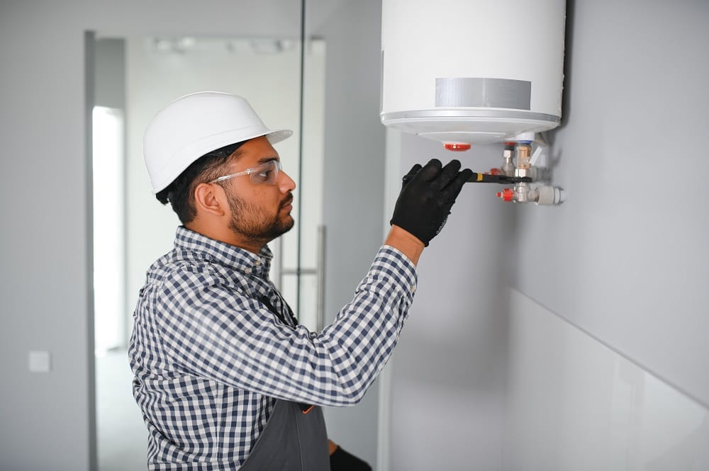 Indian plumber wearing checkered shirt and black latex gloves installing or repairing white hot water tank on grey wall