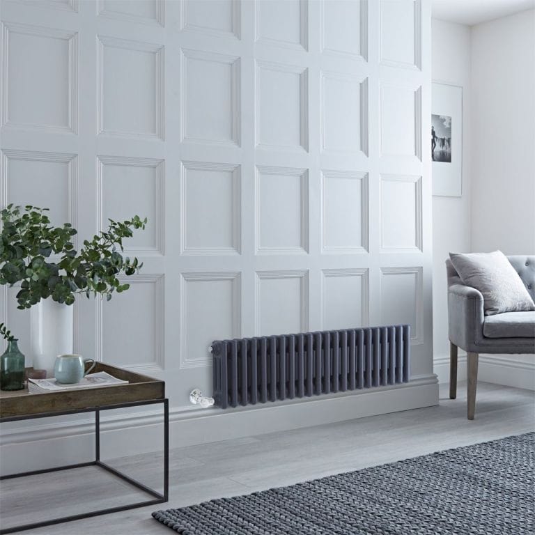 Milano Windsor anthracite traditional horizontal electric triple column radiator on white wall nearby grey chair and small brown table with white vase