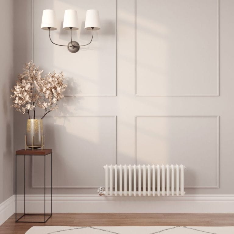 Milano Windsor white traditional horizontal electric triple column radiator on white wall nearby small table with vase and fake houseplant beneath triple light fitting