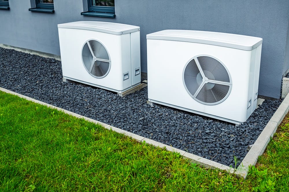 Pair of white air source heat pumps installed outside modern property on grey gravel nearby grass lawn