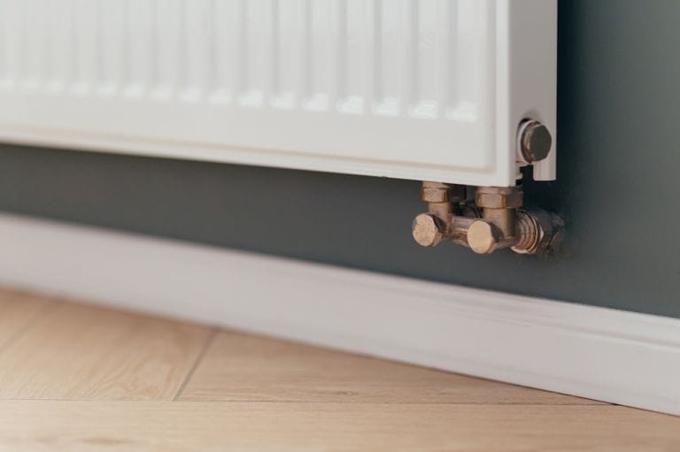 Copper radiator valves at foot of white convector radiator above skirting board and wooden effect floor