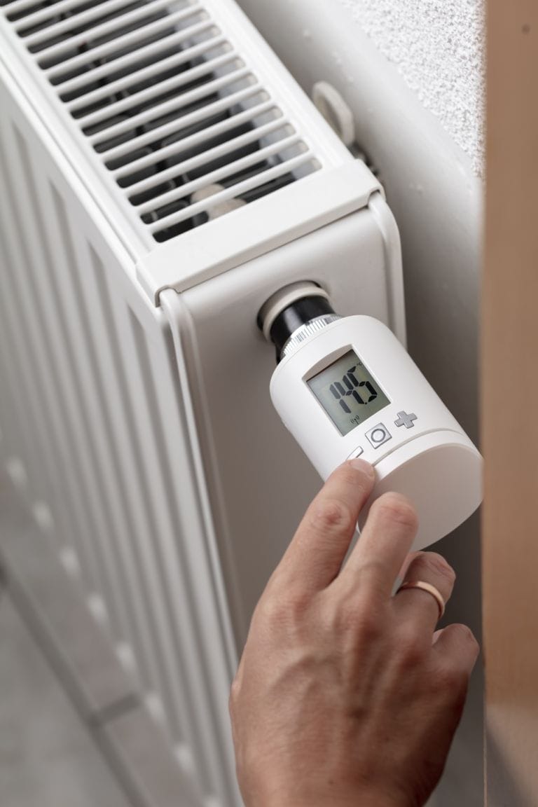 Female hand adjusting temperature on digital display thermostat valve attached to white convector radiator
