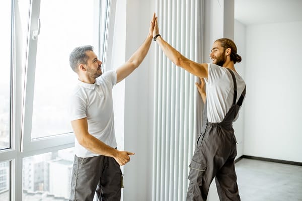 Two professional workmen in workwear installing decorative radiator in the white living room, finishing the repair in the apartment Two professional workmen in workwear installing decorative radiator in the white living room, finishing the repair in the apartment