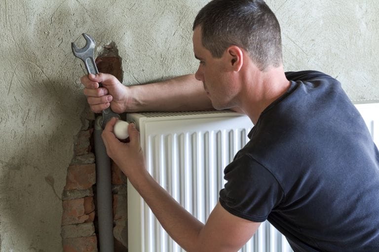 Man using spanner to reconnect a white convector radiator to wall nearby exposed brickwork